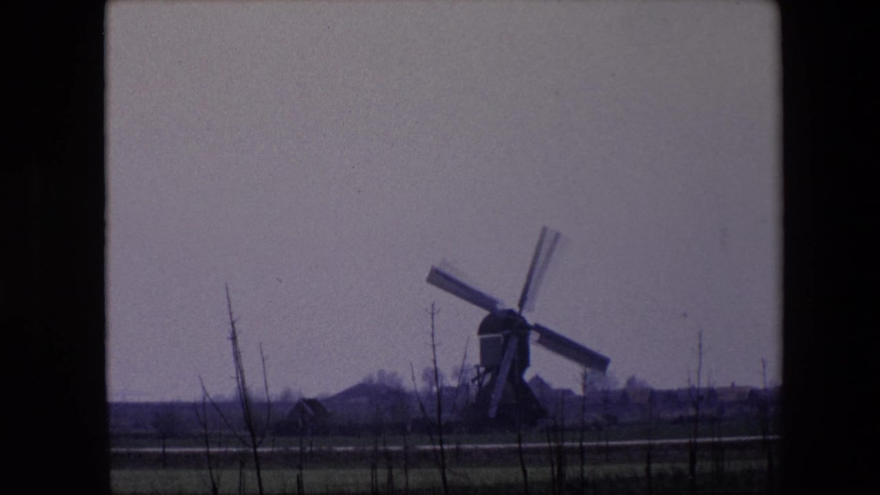 1967 Brussels-area 8mm frame showing the windmill framed to the right, with flat farmland, foreground branches, a small distant structure, and a subdued sky.