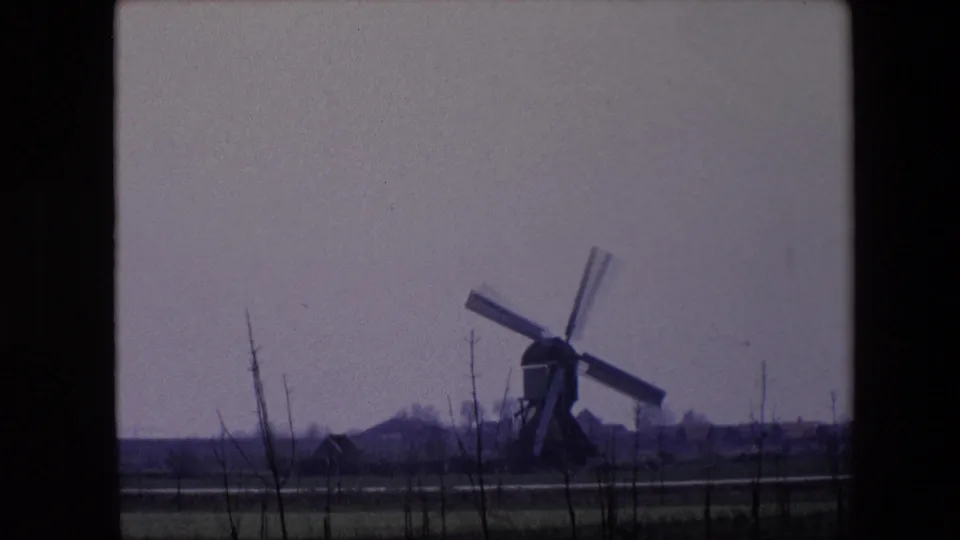 1967 Brussels-area 8mm frame showing the windmill framed to the right, with flat farmland, foreground branches, a small distant structure, and a subdued sky.