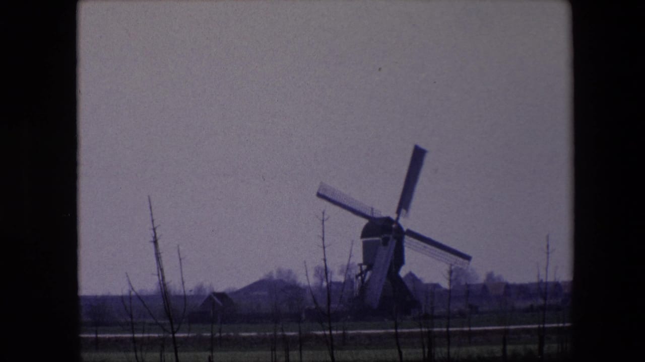 1967 8mm home-movie frame from Brussels, Belgium, showing a distant traditional windmill beyond fields, bare saplings, and low houses under an overcast sky.