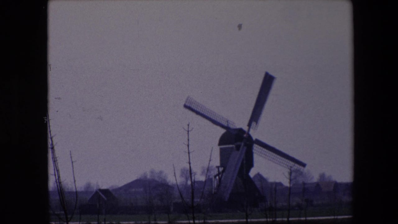 1967 Brussels-area home-movie frame showing the windmill more centered, with crossed sails, low houses, bare trees, and muted fields visible through dark frame borders.