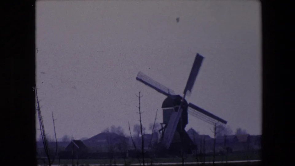 1967 Brussels-area home-movie frame showing the windmill more centered, with crossed sails, low houses, bare trees, and muted fields visible through dark frame borders.