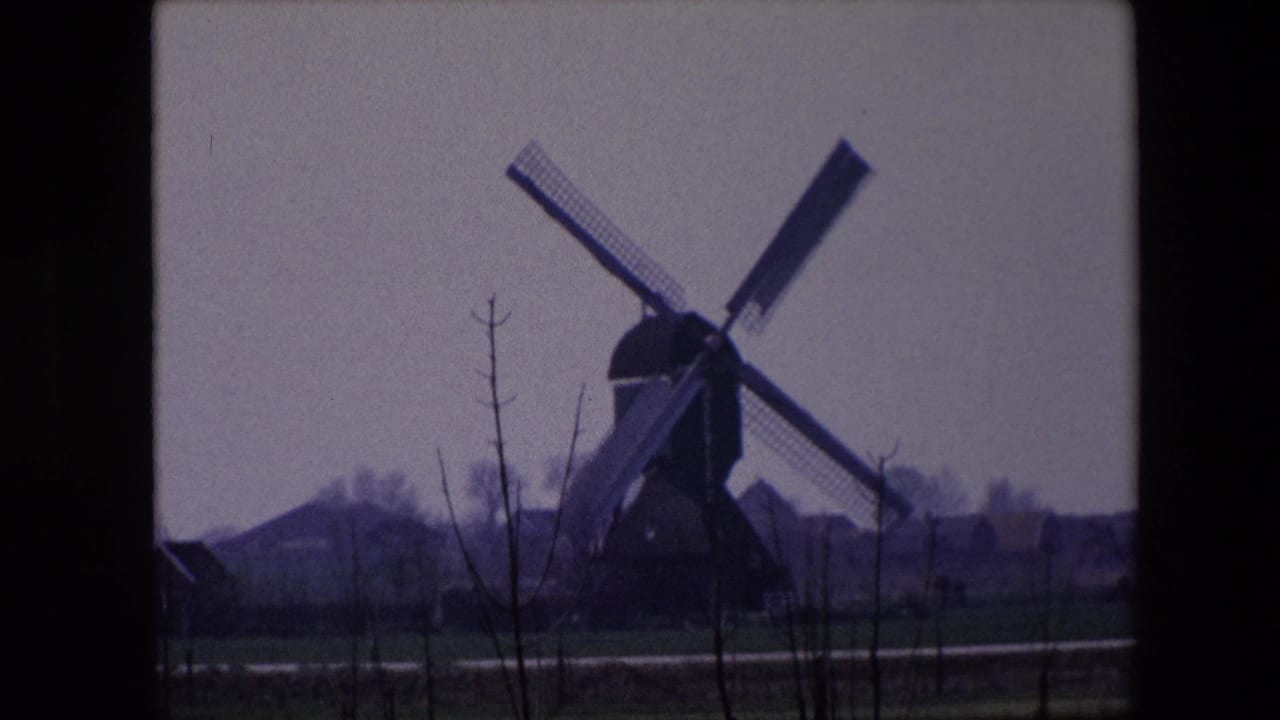 1967 Brussels home-movie frame showing the windmill at right of center, bare foreground trees, a narrow road or path, green-brown fields, and distant low buildings.
