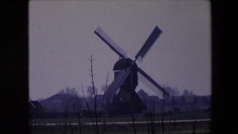 1967 Brussels home-movie frame showing the windmill at right of center, bare foreground trees, a narrow road or path, green-brown fields, and distant low buildings.