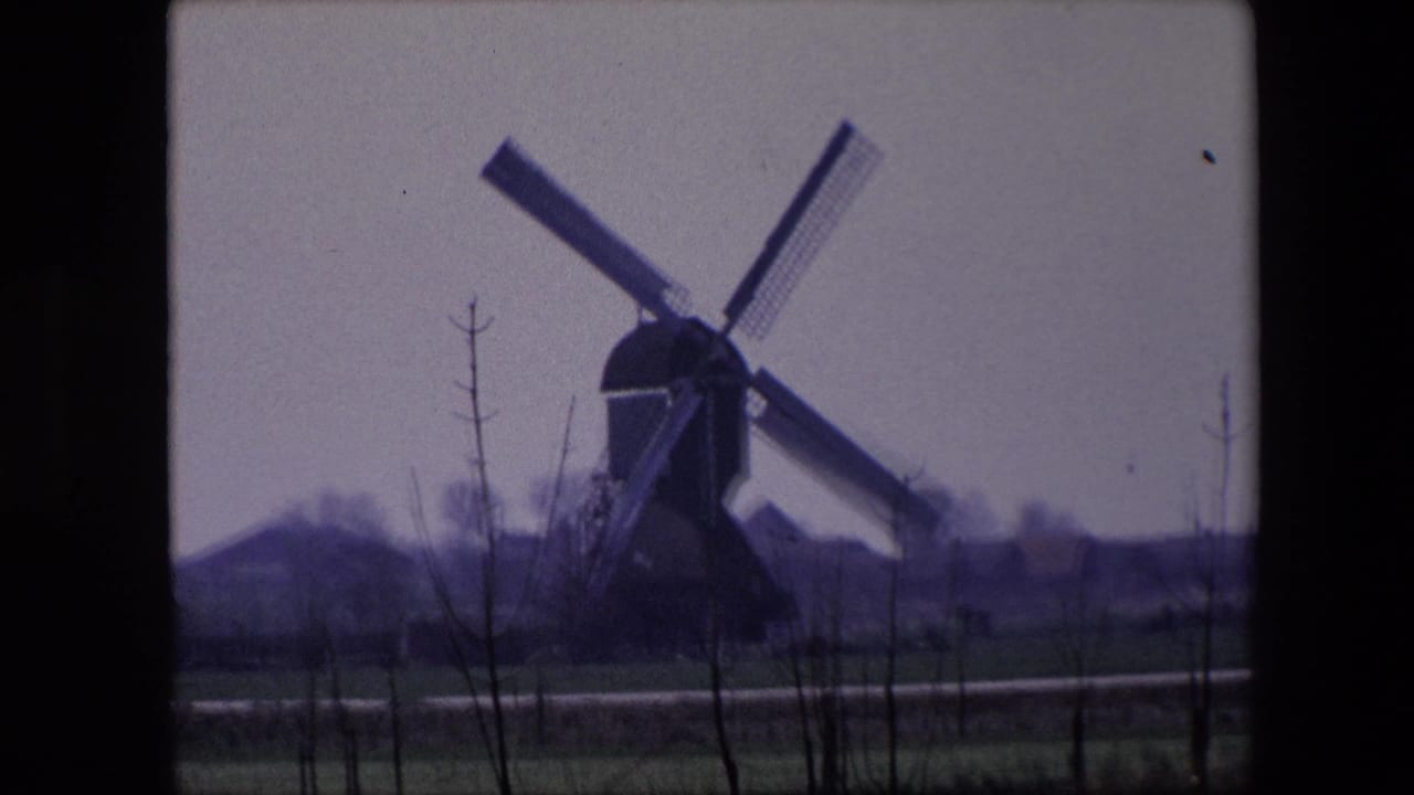 1967 8mm Brussels-area rural view of a traditional windmill in the distance, with sails angled across a pale sky and bare saplings in the foreground.