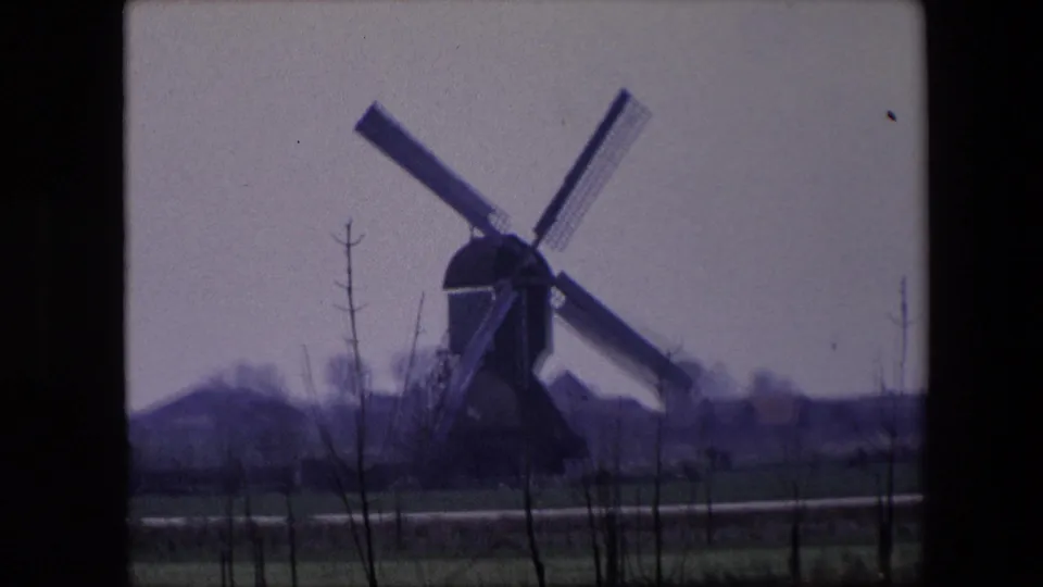 1967 8mm Brussels-area rural view of a traditional windmill in the distance, with sails angled across a pale sky and bare saplings in the foreground.