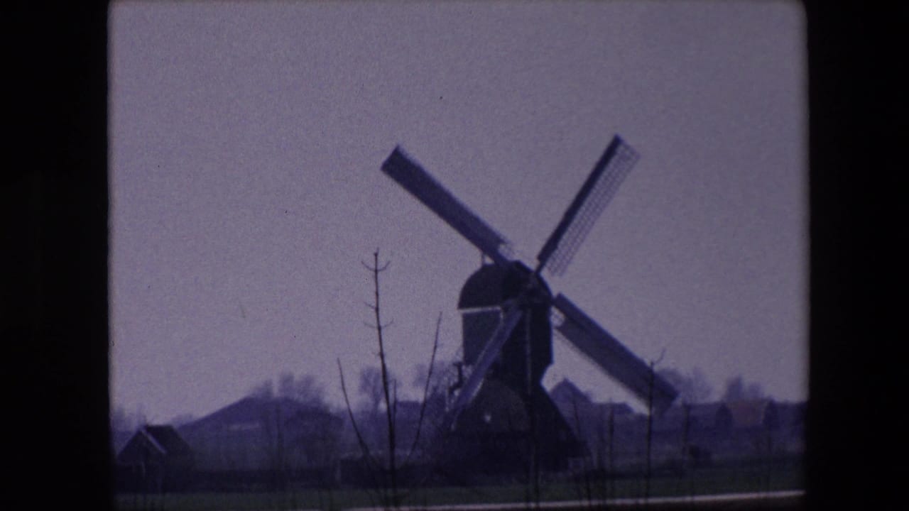 Traditional windmill near Brussels in 1967 home-movie footage, seen across open ground with bare trees and low structures under an overcast sky.