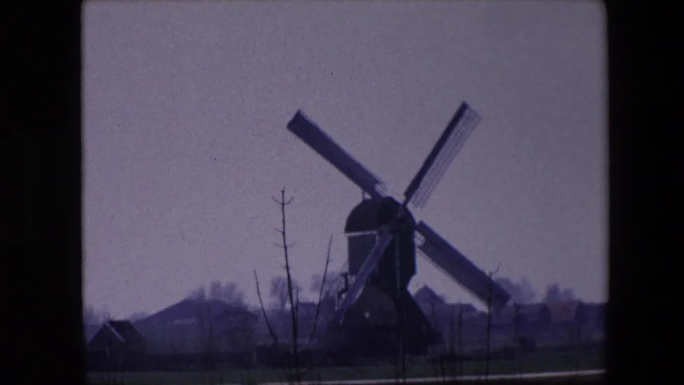 Traditional windmill near Brussels in 1967 home-movie footage, seen across open ground with bare trees and low structures under an overcast sky.