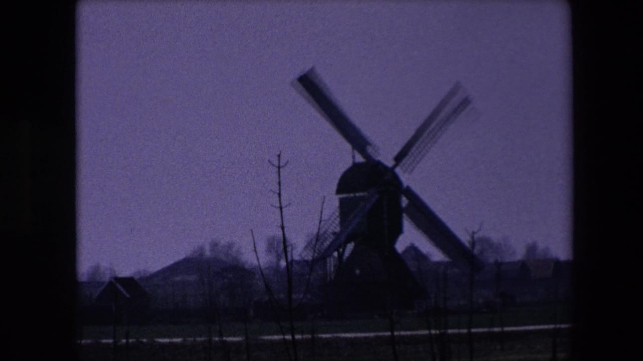 Faded 8mm frame from the 1967 Brussels-area clip showing the windmill lower in the scene, with foreground saplings, dimmer exposure, and rural buildings nearby.