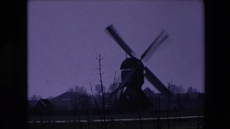 Faded 8mm frame from the 1967 Brussels-area clip showing the windmill lower in the scene, with foreground saplings, dimmer exposure, and rural buildings nearby.