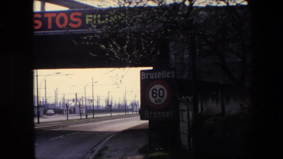 1967 Brussels highway approach beneath an overpass, showing the bilingual Bruxelles/Brussel 60 km sign, roadside poles, bare trees, and advertising on the bridge.