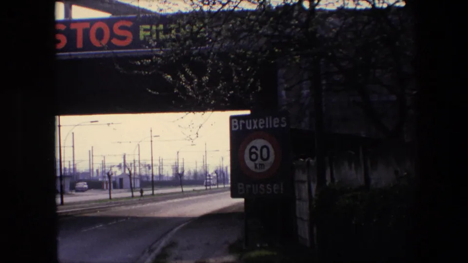 Shadowed 1967 Brussels road view with the Bruxelles/Brussel 60 km speed sign at right, open roadway at left, roadside poles, and overpass advertising above.