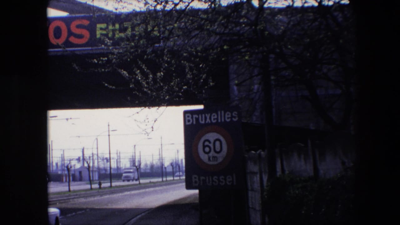 1967 Brussels home-movie frame showing a Bruxelles Brussel 60 km/h speed limit sign beside a road with classic cars, utility poles, bare trees, and a dark foreground frame.