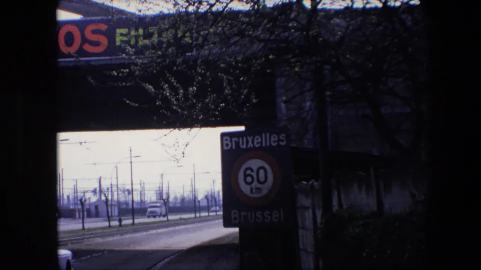 1967 Brussels home-movie frame showing a Bruxelles Brussel 60 km/h speed limit sign beside a road with classic cars, utility poles, bare trees, and a dark foreground frame.