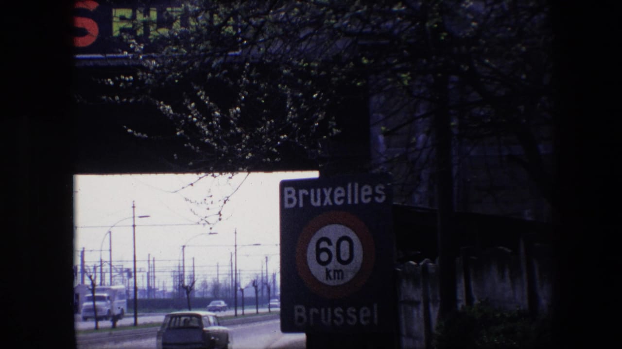 1967 Brussels frame with the bilingual Bruxelles Brussel 60 km/h sign close to the camera, classic cars on the street, roadside poles, bare branches, and a dark upper foreground.