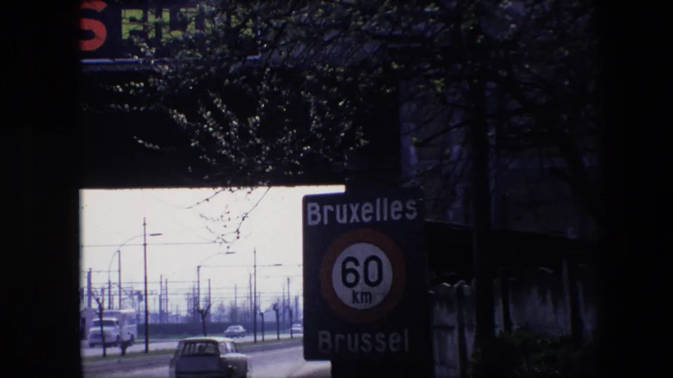 1967 Brussels frame with the bilingual Bruxelles Brussel 60 km/h sign close to the camera, classic cars on the street, roadside poles, bare branches, and a dark upper foreground.
