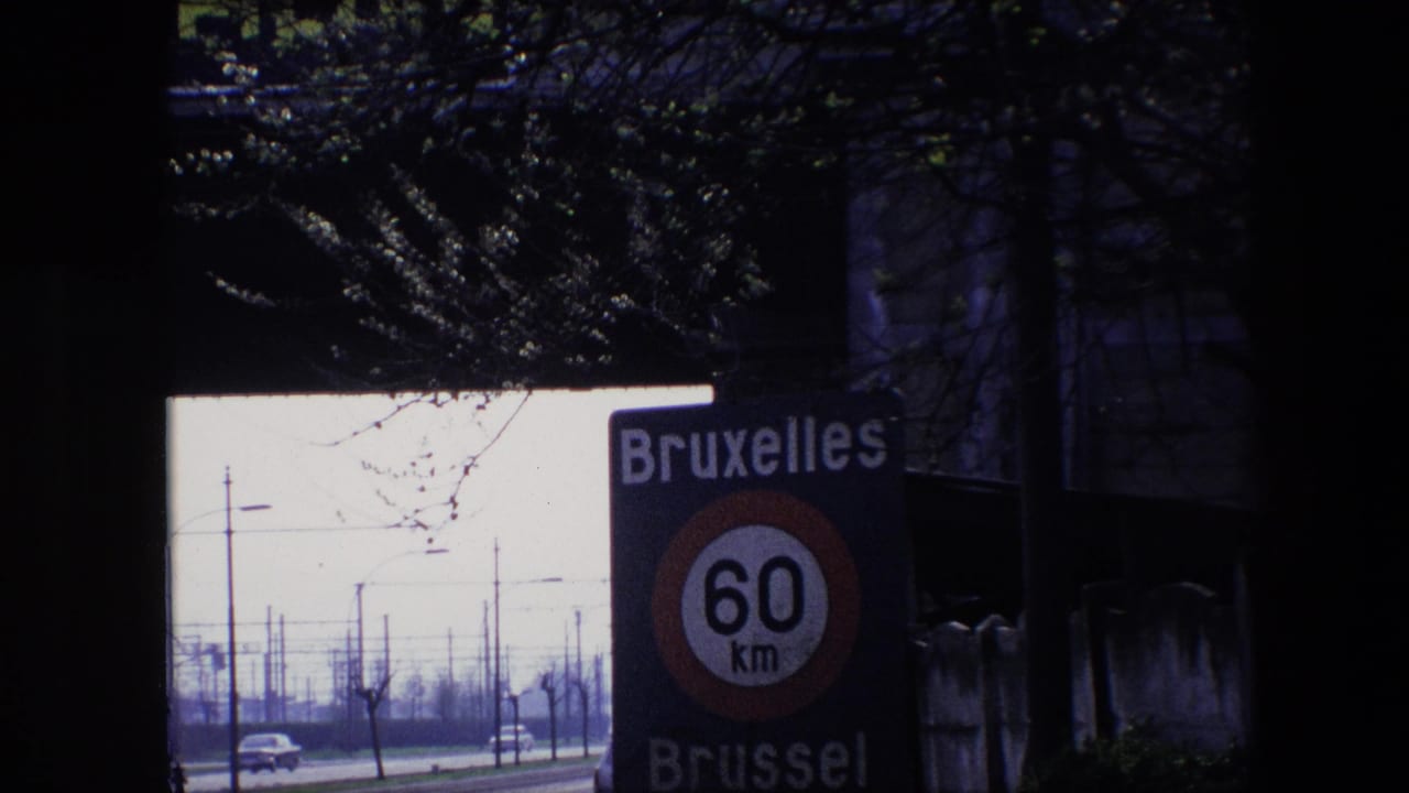 1967 Brussels 8mm frame showing a closer view of the Bruxelles Brussel 60 km/h road sign, with classic cars, poles, and bare roadside trees visible through a dark opening.