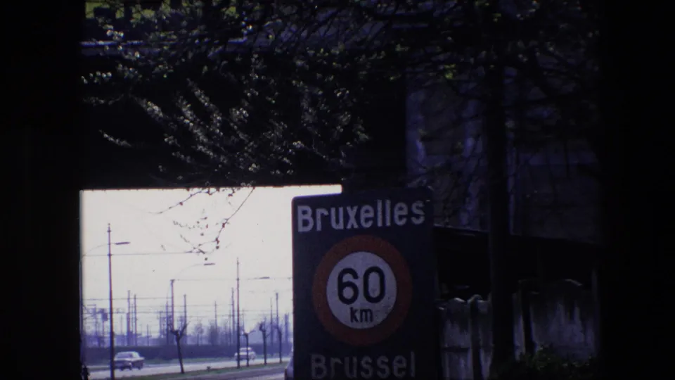 1967 Brussels 8mm frame showing a closer view of the Bruxelles Brussel 60 km/h road sign, with classic cars, poles, and bare roadside trees visible through a dark opening.