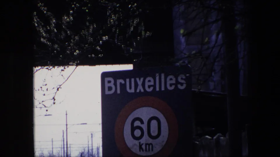 1967 Brussels footage showing a blue Bruxelles road sign and 60 km/h speed-limit roundel seen through dark tree branches against a pale sky.