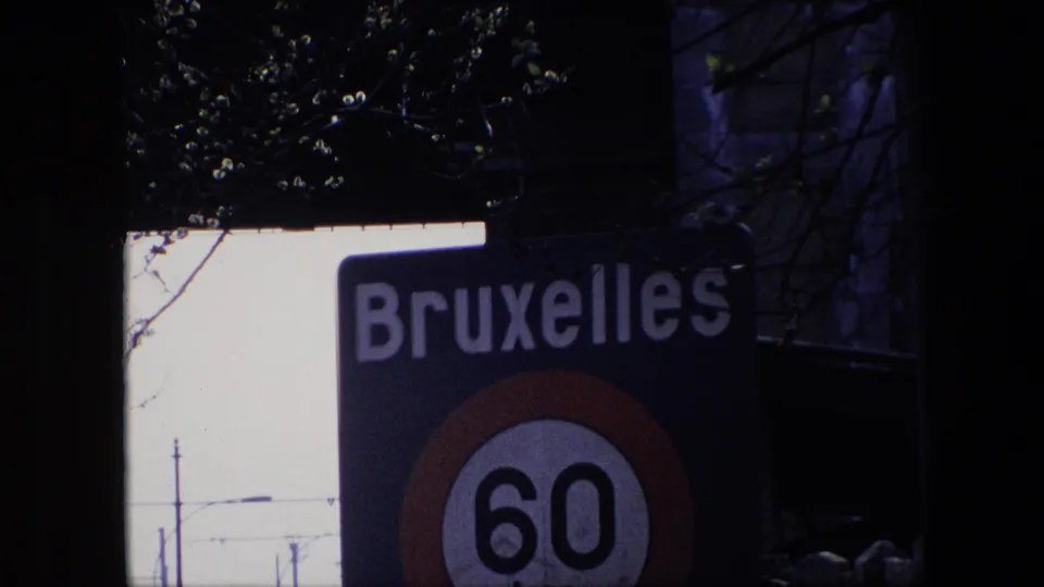 Cropped 1967 Brussels frame with the word Bruxelles visible above a partly cut off 60 km/h speed-limit sign, with branches and dark foreground shapes around it.