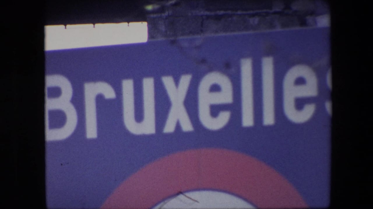 1967 Brussels home-movie frame showing a tightly cropped blue Bruxelles sign with large white letters, part of a red-and-white circular emblem, and visible 8mm frame edge shadows.
