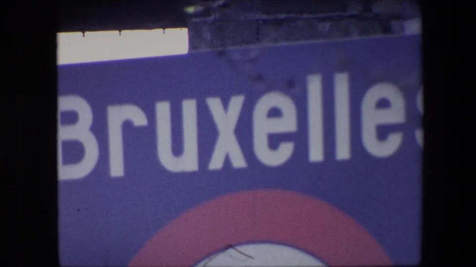 1967 Brussels home-movie frame showing a tightly cropped blue Bruxelles sign with large white letters, part of a red-and-white circular emblem, and visible 8mm frame edge shadows.