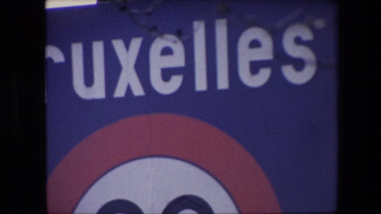 1967 Brussels frame with a cropped public sign reading part of Bruxelles, a red-and-white circular emblem below, and soft focus from the 8mm home-movie scan.