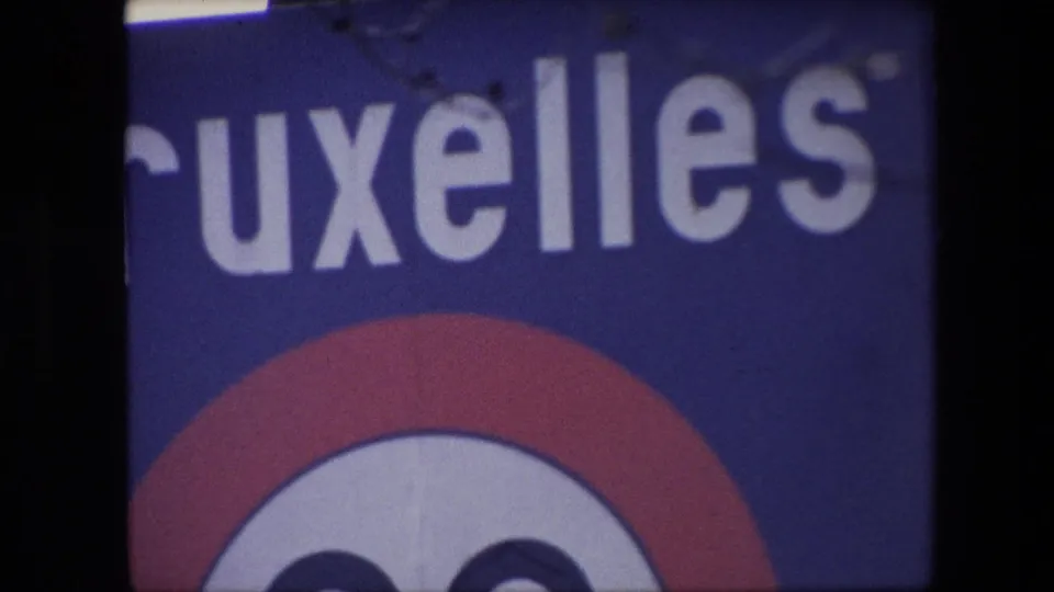 1967 Brussels frame with a cropped public sign reading part of Bruxelles, a red-and-white circular emblem below, and soft focus from the 8mm home-movie scan.