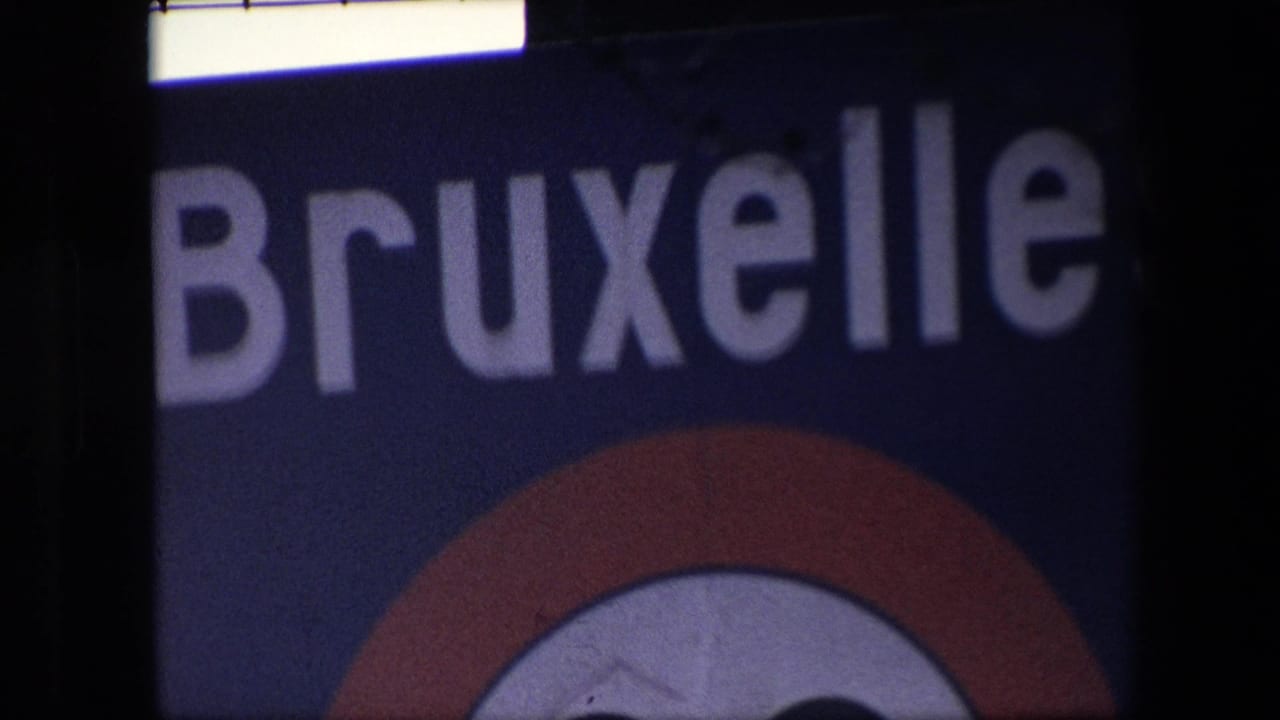 Dim 1967 Brussels home-movie frame showing a close, underexposed crop of the Bruxelles lettering and lower red circular emblem within heavy film borders.