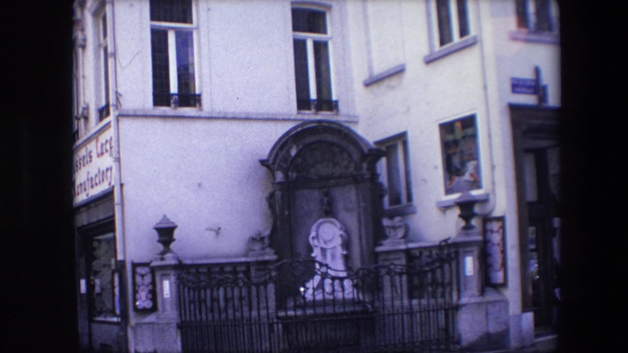 1967 Brussels street-level frame showing a pale building facade with arched windows, a dark framed monument or niche, black iron fencing, and a small statue-like figure near the base.