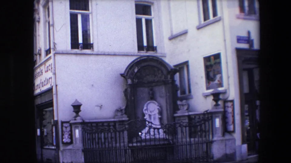 1967 Brussels street-level frame showing a pale building facade with arched windows, a dark framed monument or niche, black iron fencing, and a small statue-like figure near the base.
