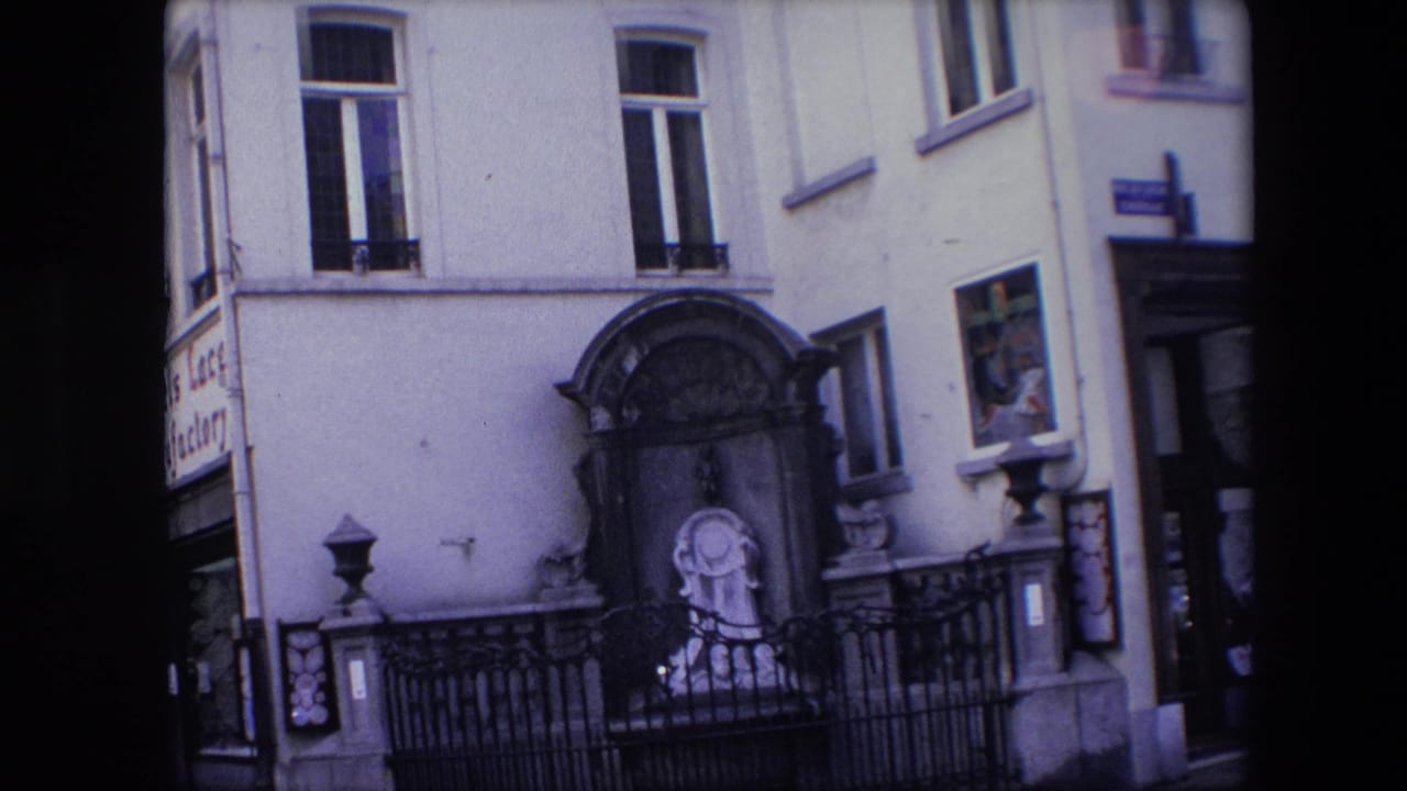 1967 Brussels home-movie frame showing an arched stone shrine with a religious statue, iron railing, tall windows, and partial commercial signage at the edge of the facade.