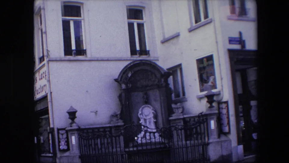 1967 Brussels home-movie frame showing an arched stone shrine with a religious statue, iron railing, tall windows, and partial commercial signage at the edge of the facade.