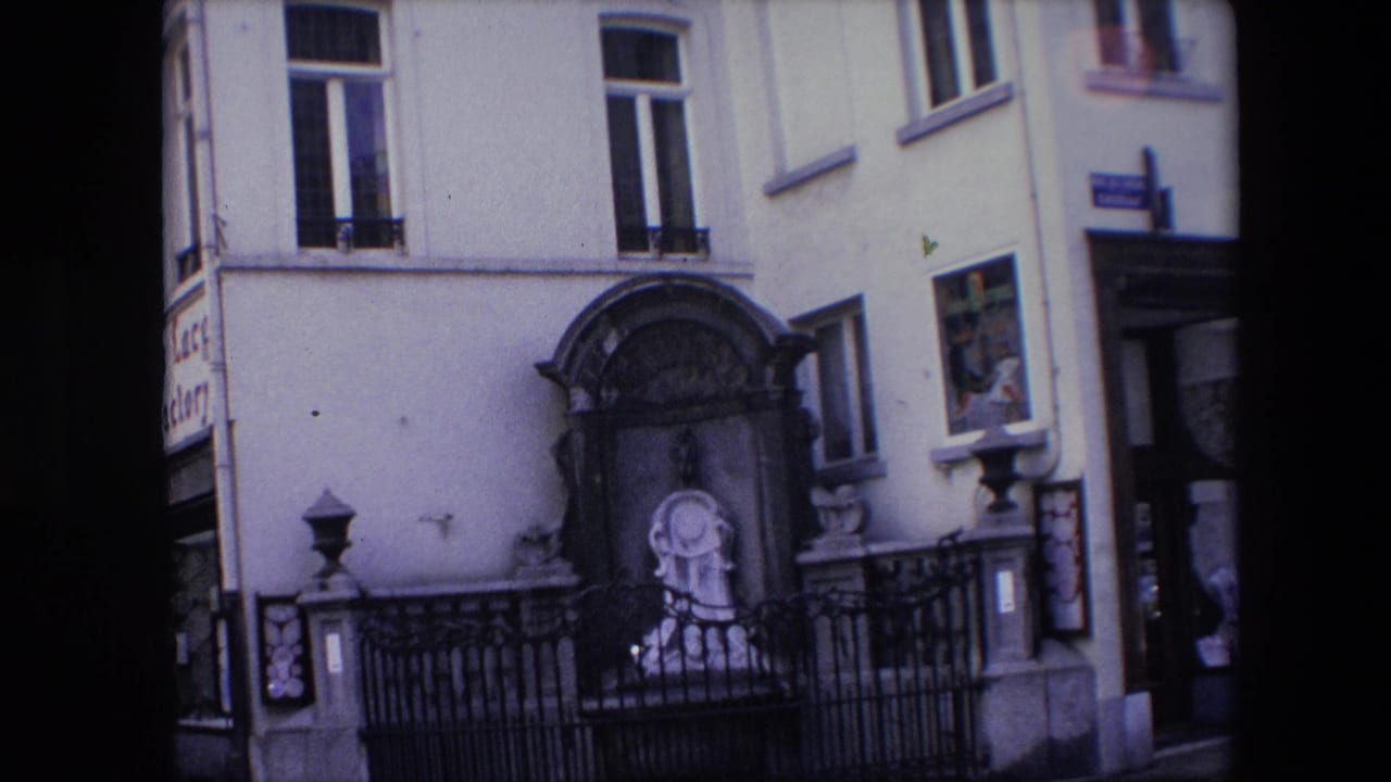 1967 Brussels frame with the shrine centered against a pale facade, bordered by black iron railings, upper windows, a side doorway, and a partially visible sign.