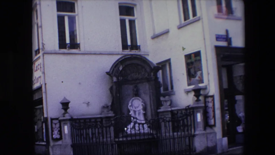 1967 Brussels frame with the shrine centered against a pale facade, bordered by black iron railings, upper windows, a side doorway, and a partially visible sign.