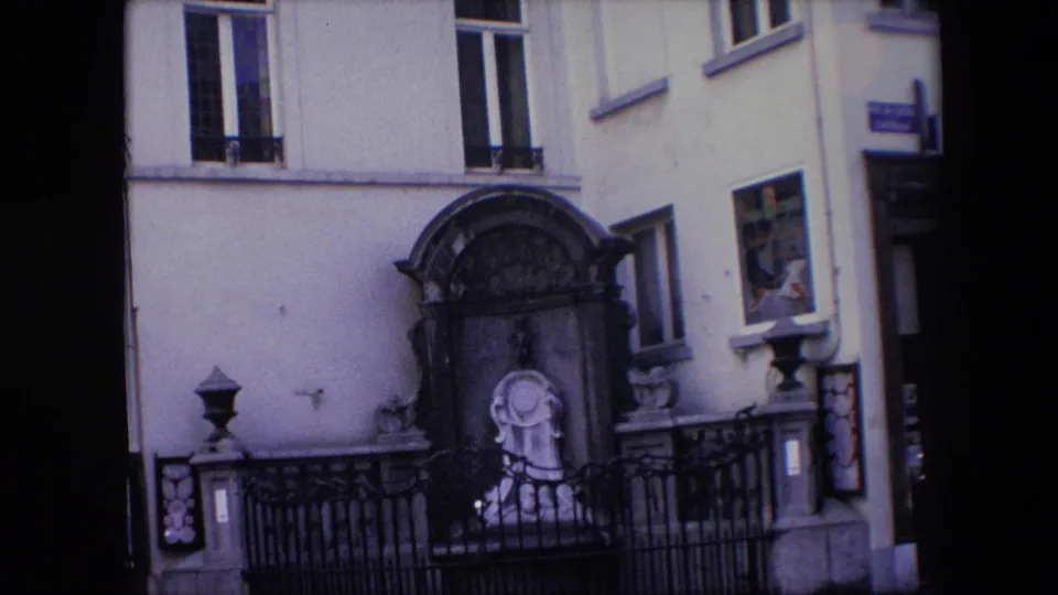1967 Brussels 8mm frame showing a closer handheld view of the facade shrine, dark iron fence, tall windows, and a partial Lace Factory sign along the left edge.