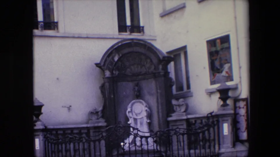 1967 Brussels frame showing the shrine and statue slightly closer and lower in the composition, with iron railings, pale wall, tall windows, and adjacent doorway details.