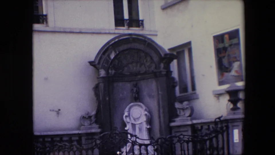 1967 Brussels home-movie frame showing an ornate stone fountain with a small child-like statue, carved basin detail, iron railing, and pale building facade.