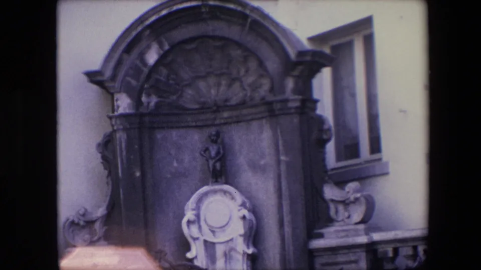 Zoomed 1967 Brussels home-movie frame emphasizing the fountain's dark stone arch, small central figure, pale carved basin form, and flanking ornament.