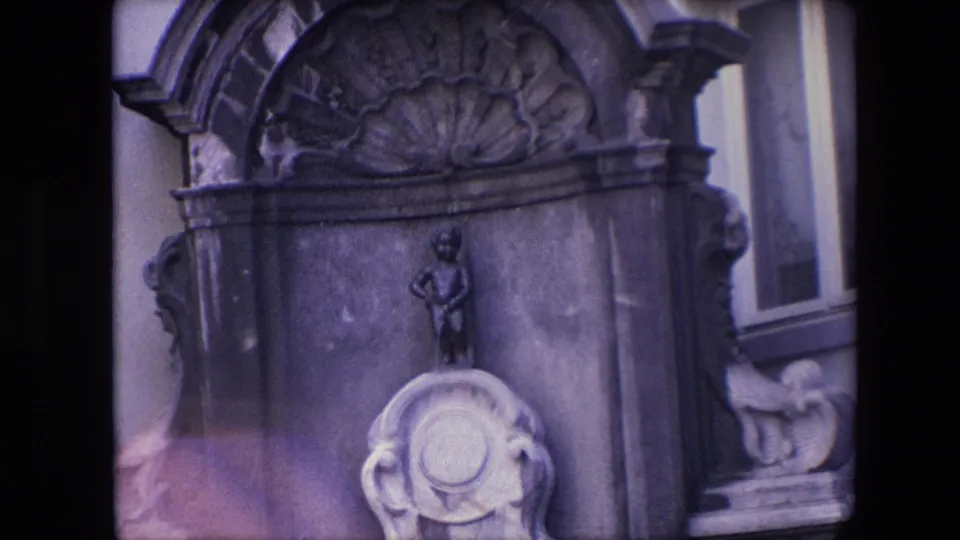 Tight 1967 Brussels home-movie frame showing the ornate fountain top, small child-like statue, pale carved basin, side ornament, window, and facade edge.