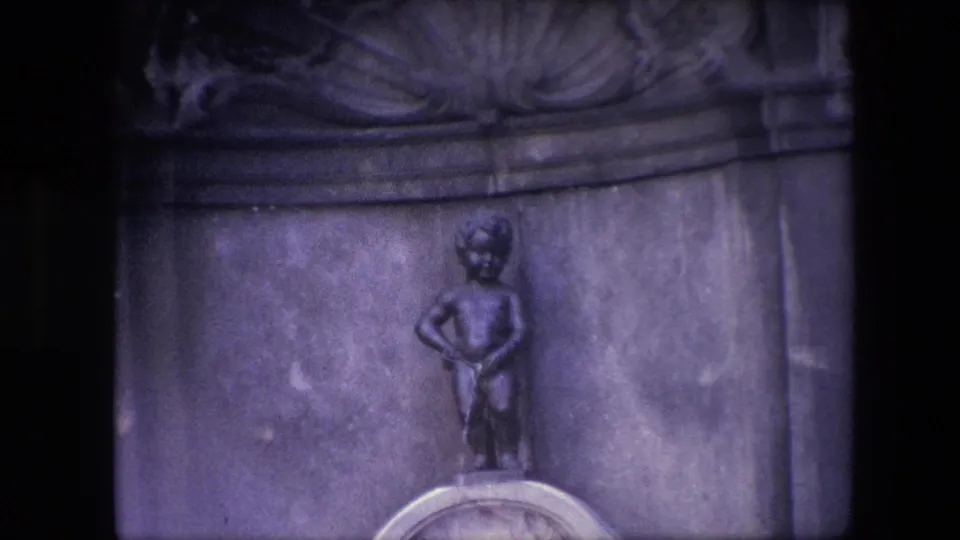 A tighter 1967 Brussels home-movie view shows the statue against the stone backdrop, with the rounded pedestal partly cropped by the frame.