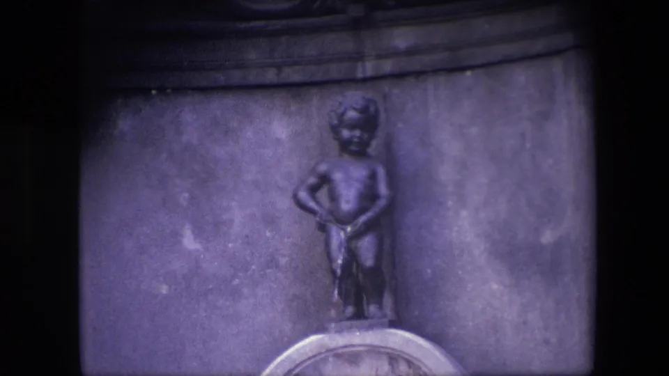 Final 8mm frame holds on the Manneken Pis figure against the muted stone wall, with the rounded pedestal detail visible along the lower edge.