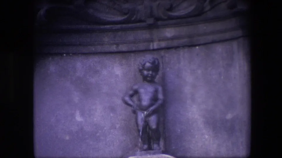 1967 Brussels home-movie frame with the Manneken Pis statue slightly lower in view, showing more of the stone basin rim and dark background detail.