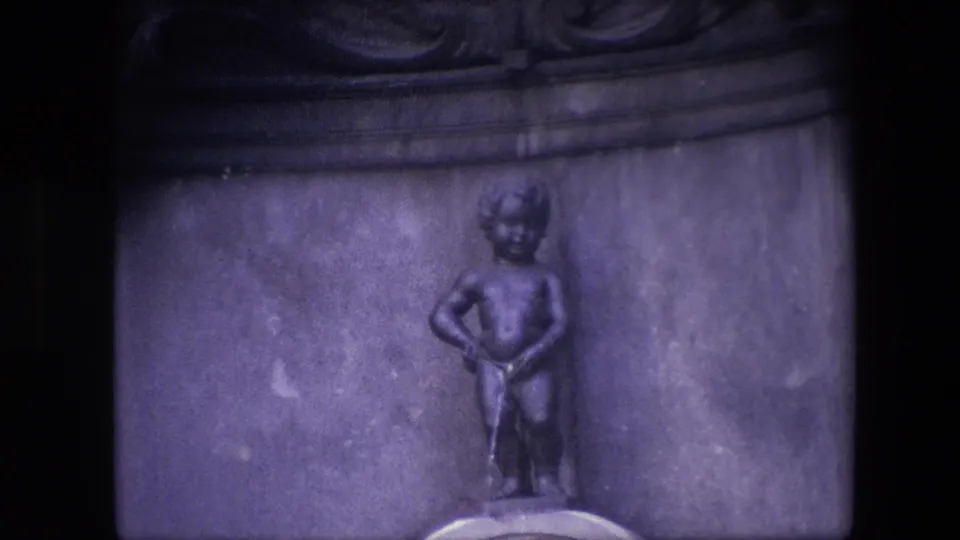 Close 1967 Brussels home-movie view of the Manneken Pis bronze figure set into a stone fountain niche with dark film borders and a purple cast.