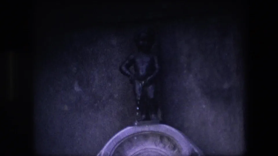 A dimmer 8mm frame shows the small bronze Manneken Pis figure against the stone wall, with the fountain basin edge at the bottom and heavy exposure falloff.
