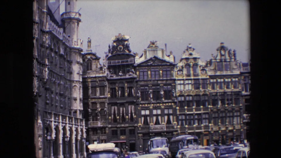 1967 Brussels street view from 8mm footage with decorative rooflines, multi-story facades, parked mid-century cars, and a bus near the curb.