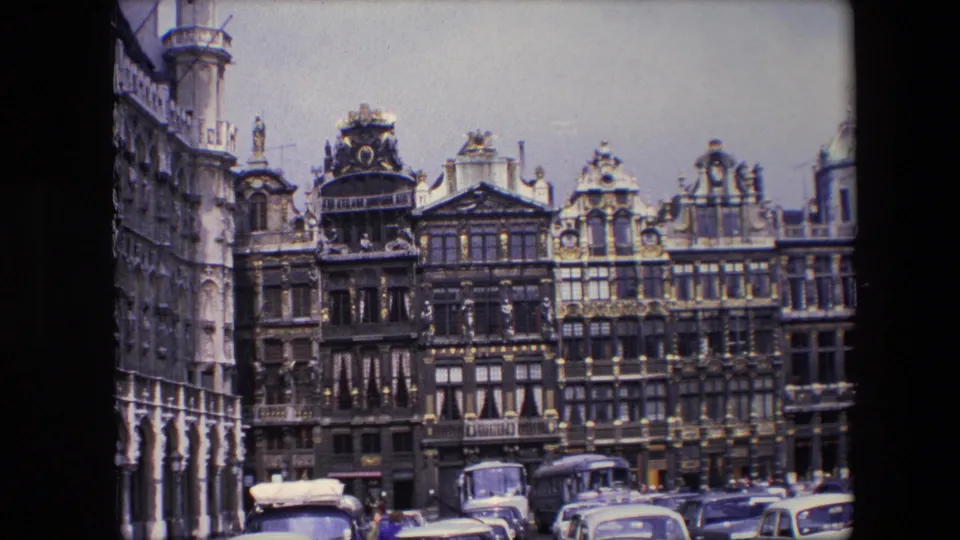 1967 Brussels home-movie frame showing ornate stone and brick facades, arcaded architecture at left, parked automobiles, and small pedestrians near the street.