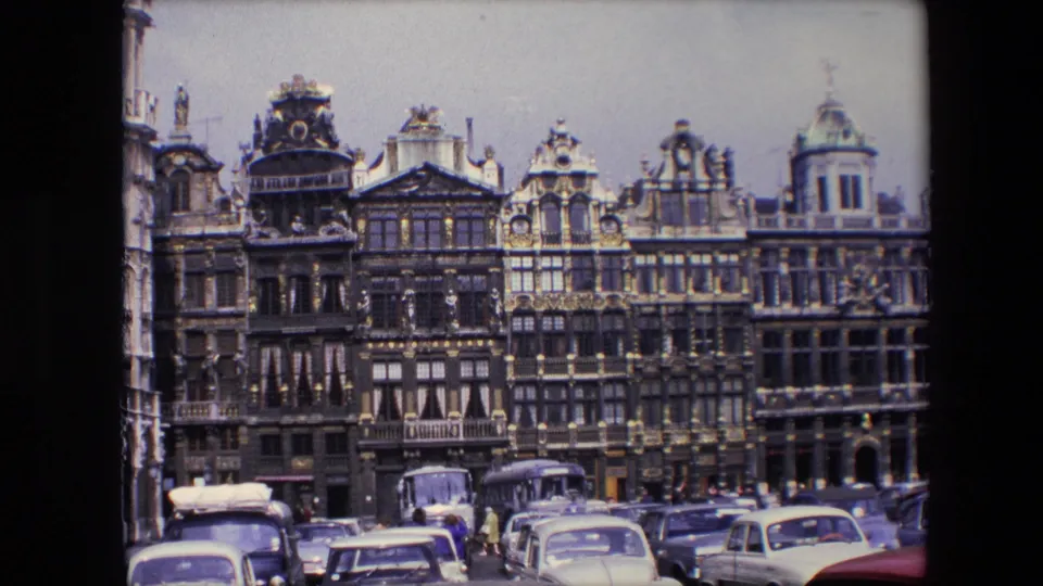 1967 Brussels travel footage frame with richly decorated Grand Place buildings, parked cars, buses, and small pedestrians in the square foreground.