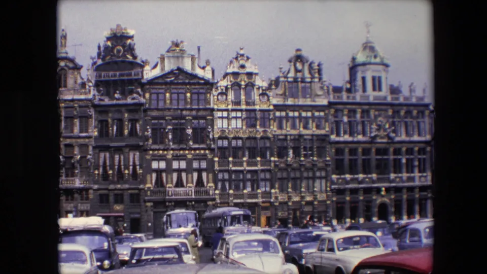 A 1967 8mm frame from Brussels showing Grand Place architecture with layered gables, parked European cars, a bus, and distant pedestrians.