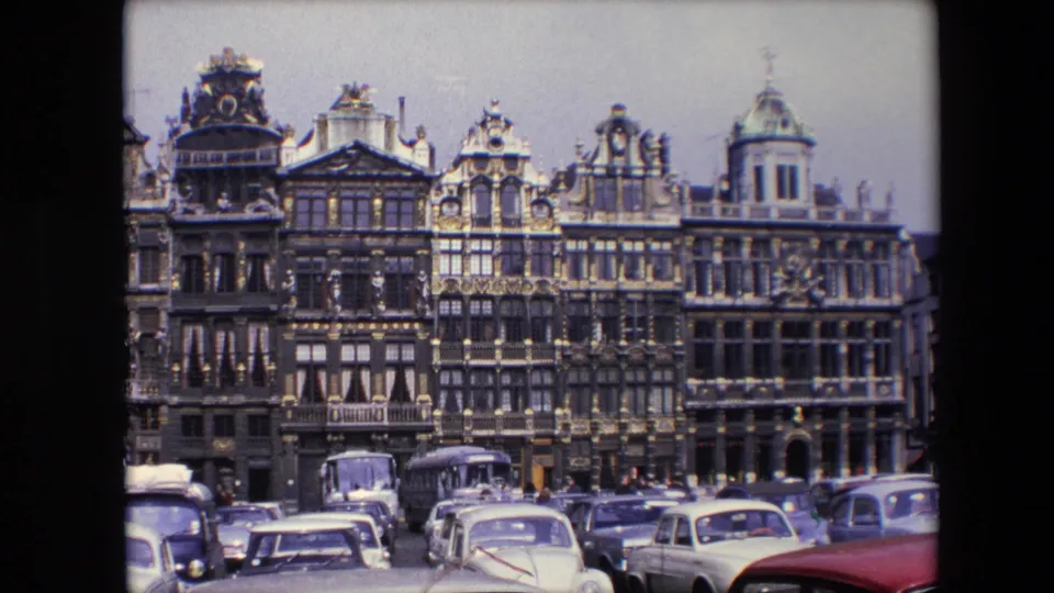 1967 Brussels, Belgium frame showing decorated Grand Place facades, cars and buses in the foreground, and distant pedestrians in a public square.