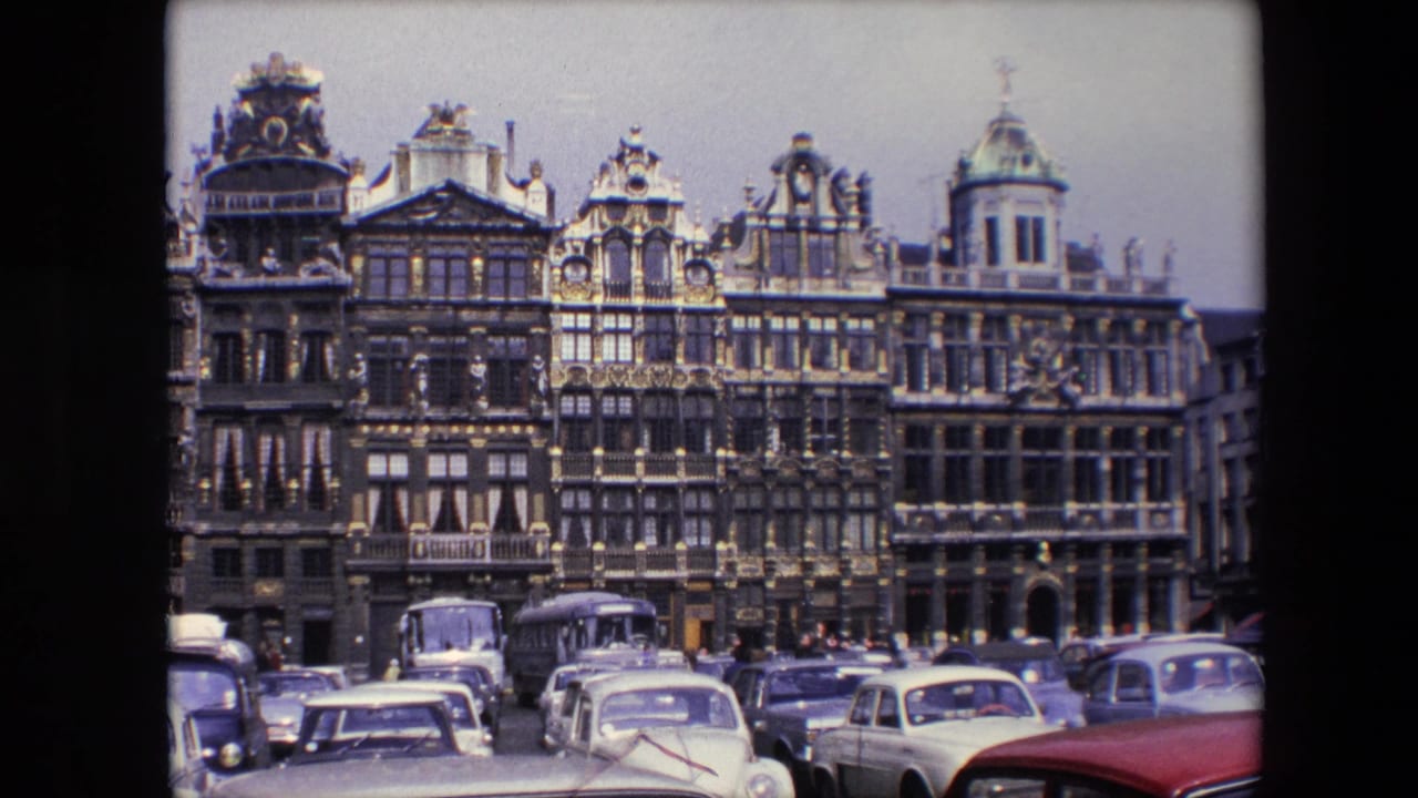 1967 Brussels home-movie frame showing ornate historic facades with gold-toned detailing above a foreground of parked cars and street traffic.